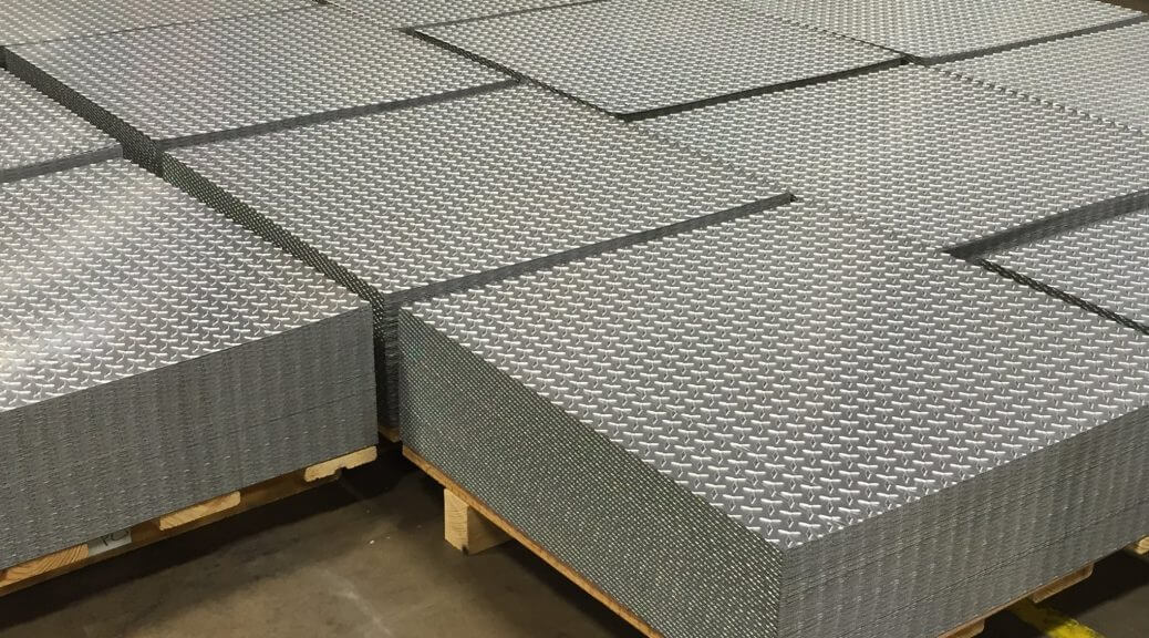 Stacked textured metal diamond-plate sheets on wooden pallets in a warehouse, gray tones.